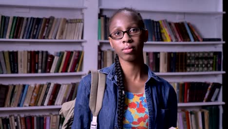 Young-pretty-african-american-woman-in-glasses-reading-book-in-library,-looking-at-camera,-serious-and-concentrated,-bookshelves-background