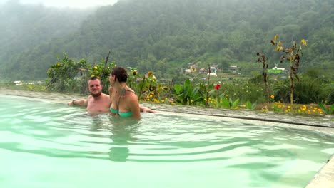 Ein-Mann-sitzt-in-einem-Pool-mit-Thermalwasser.-Eine-Frau-betritt-den-Pool-und-küsst-einen-Mann