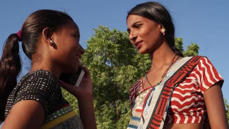 Tiro-bajo-mano-de-las-mujeres-hablando-de-comunicación-de-dispositivo-móvil-del-teléfono-celular-mientras-amigo-interesado-maquillaje-de-joyería-de-traje-de-curioso-interés-feliz-alegre-lindo-mano-sol-verano-sari-caliente-al-aire-libre-Vestido