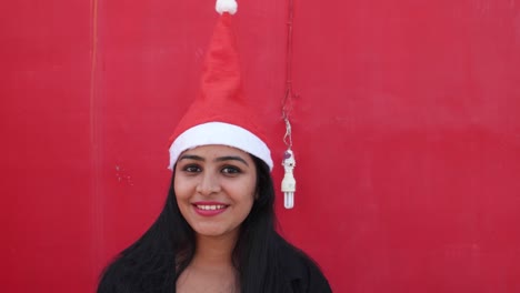 Young-Indian-woman-with-Santa's-hat-welcome-and-making-hand-gestures,-excited-and-super-happy,-with-a-matching-red-background,-handheld-gimbal-stabilized