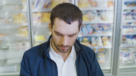 Portrait-Shot-of-the-Handsome-Man-Choosing--Tin-Can-from-the-Canned-Goods-Section-and-Places-it-In-His-Shopping-Cart.-In-the-Background-Frozen-Goods-Section-of-the-Store.