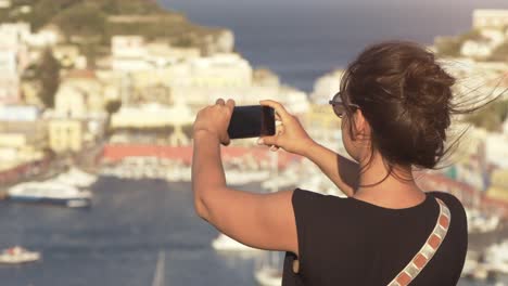 Happy-young-woman-tourist-taking-smartphone-pictures-of-town-on-Ponza-island-in-Italy-traditional-sea-city-landscape