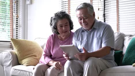 Senior-pareja-sujetando-el-tablet-y-sentados-en-el-sofá-juntos-en-la-sala-de-estar-en-casa.-Concepto-de-familia-feliz,-post-jubilación,-calidad-vida,-salud-mental-y-psicología-positiva.-resolución-de-4-k.