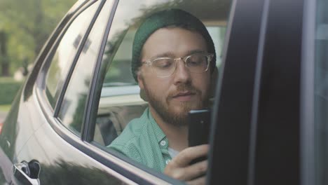 Stilvolle-junge-Mann-fahren-in-einem-Auto-sitzen-auf-dem-Beifahrer-Sitz-nutzt-Smartphone-Typen-Nachricht,-blättert-durch-Internet.-Kamera-Schuss-von-außerhalb-des-Fahrzeugs.