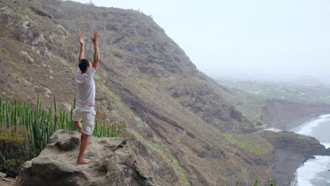 Forma-joven-practica-yoga-de-saludo-al-sol-en-la-montaña-por-mar.-Joven-disfrutando-de-la-meditación-y-el-yoga.-concepto-de-fitness,-deporte,-gente-y-estilo-de-vida---hombre-joven-haciendo-ejercicios-de-yoga