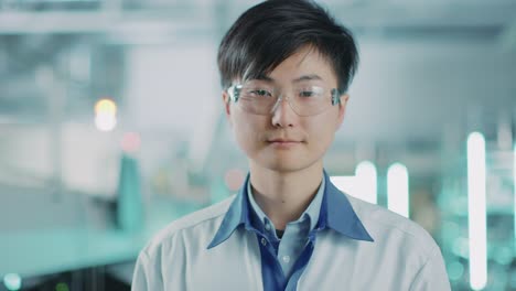 On-High-Tech-Factory:-Portrait-of-Asian-Worker-Wearing-Uniform-and-Safety-Goggles.-In-the-Background-Blurred-Electronics-Assembly-Line-with-Bright-Lights-and-other-Workers-Doing-their-Jobs.