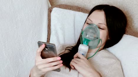 Verwenden-Sie-Vernebler-und-Inhalator-für-die-Behandlung.-Junge-Frau-Einatmen-durch-Inhalator-Maske-auf-der-Couch-liegen-und-im-Handy-Chat.-Seitenansicht.