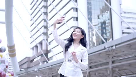 Young-Asian-businesswoman-Using-Smart-Phone-are-taking-selfie,-smiling-happily-in-city-centre-Bangkok,-Thailand.-everyday-life-Technology-Concept.-SLOW-MOTION