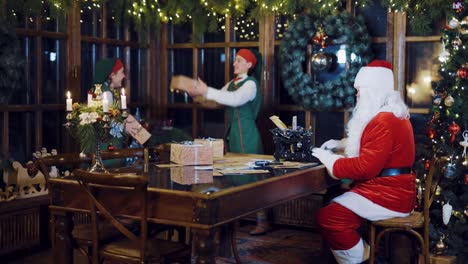 Santa-Claus-is-writing-a-letter-on-the-typewriter-and-watching-as-the-Elves-are-throwing-each-other-presents-on-the-background-of-Christmas-decorations-in-the-room-on-the-eve-of-the-New-Year