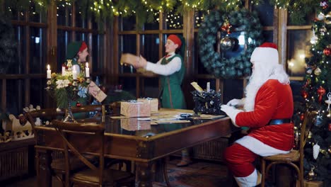 Santa-Claus-está-escribiendo-una-carta-en-la-máquina-de-escribir-y-viendo-como-los-elfos-están-tirando-unos-a-otros-presentes-en-el-fondo-de-adornos-de-Navidad-en-la-sala-en-la-víspera-del-año-nuevo