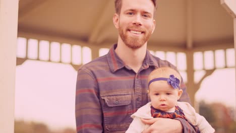 Portrait-of-a-young-man-sitting-with-his-baby-daughter-under-a-gazebo-on-a-fall-day