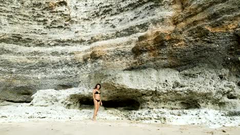 Una-chica-en-bañador-en-la-playa.-Contra-el-fondo-de-un-enorme-muro-de-piedra.-Ella-camina-a-lo-largo-de-la-playa.-Camina-hacia-adelante-y-hacia-atrás.-Bali