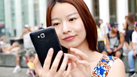 Mujer-asiática-atractiva-usando-el-teléfono-en-la-ciudad.-Ojos-oscuros-y-cabello-oscuro.-Fondo-lleno-de-gente-y-urbano.