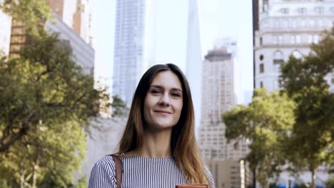 Portrait-of-young-happy-businesswoman-standing-in-the-park-of-financial-centre-of-New-York,-USA-and-holding-documents