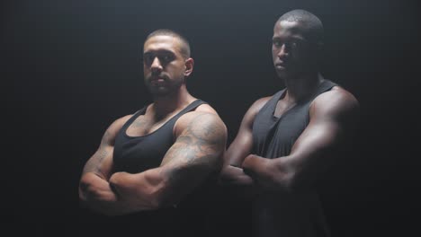 Portrait-shot-of-two-muscular-black-men-staring-at-the-camera-with-their-arms-crossed-on-a-dark-background.