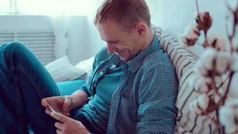 Close-up-shot-of-a-happy-young-man-sitting-on-the-couch-while-texting-on-the-smartphone-and-looking-to-the-camera,-smiling-happily