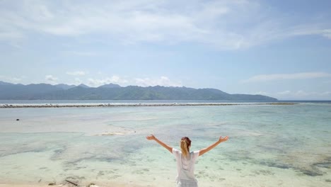 AERIAL-SHOT.-Woman-standing-by-stunning-beach-in-Bali-arms-wide-open.-Drone-flying-above-stunning-beach