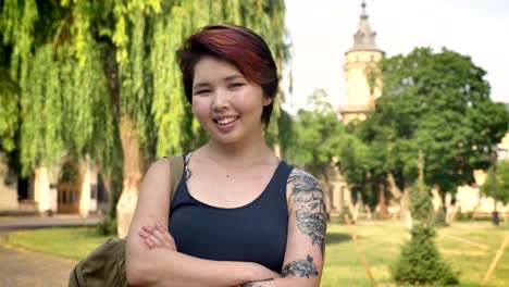 Portrait-of-young-asian-women-with-tattoo-standing-with-crossed-hands-and-looking-into-camera-in-park-near-university,-smiling,-happy