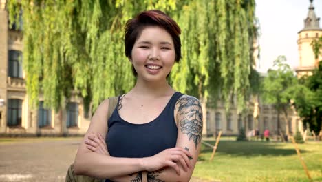 Joven-asiática-tatuada-estudiante-permanente-en-el-parque-cerca-de-la-Universidad-y-en-la-cámara,-mirando-sonriente-y-alegre