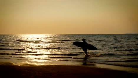 4-K.-Silhouette-des-jungen-glücklich-Surf-Mann-läuft-mit-langen-Surfbretter-bei-Sonnenuntergang-am-tropischen-Strand.-Surfer-am-Strand-im-Meer-bei-Sonnenuntergang-mit-schönen-Sonnenlicht-reflektieren-auf-der-Wasseroberfläche.