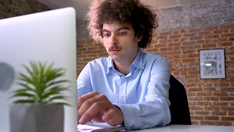 Funny-nerd-programmer-with-curly-hair-coding-on-laptop-and-sitting-at-table-in-modern-office