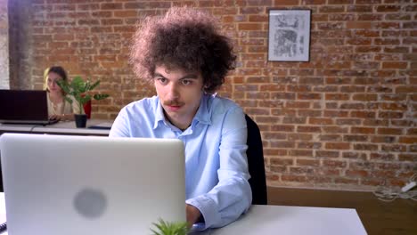 Funny-nerd-programmer-with-mustache-and-curly-hair-coding-on-laptop-sitting-at-table,-modern-office-background