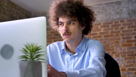 Funny-nerdy-man-with-curly-volume-hair-thinking-and-typing-on-laptop,-sitting-in-modern-office,-concentrated-and-determined