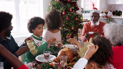 Gemischte-Rassen,-Multi-Generationen-Familie-sitzt-am-Tisch-ihre-Weihnachten-Witze-lesen-und-setzen-auf-Papier-Partyhüte-von-Christmas-Cracker,-Nahaufnahme