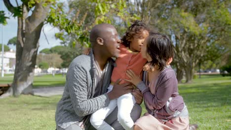 Mixed-race-daughter-hugging-and-kissing-parents-in-park