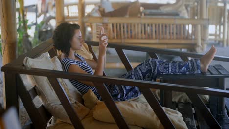 Activities-on-the-island-of-Bali.-A-girl-with-short-hair-in-a-striped-T-shirt-and-baggy-trousers-sitting-in-an-easy-chair,-put-his-feet-on-the-table.-It-relaxes-and-relieves-beautiful-views-on-your-phone