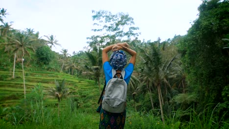 Russian-female-traveler-dressed-in-casual-blue-clothing-is-standing-in-the-middle-of-Indonesian-endless-fields,-rising-her-hands-and-enjoying-luxuriant-tropical-vegetation-and-fresh-air-around