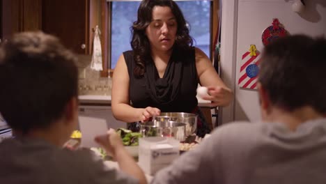 A-woman-cracks-an-egg-into-a-bowl-and-whisks-it,-her-sons-sitting-at-the-counter