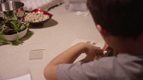 A-little-boy-puts-a-Christmas-sticker-on-a-piece-of-paper,-while-his-mother-prepares-food