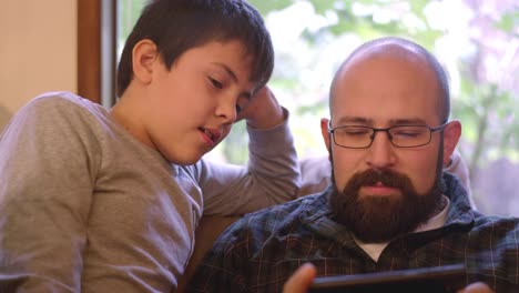 A-father-and-son-sit-and-talk-on-a-couch-at-home-while-the-dad-uses-his-phone