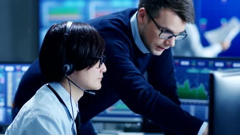 In-the-Network-Operations-Center-Senior-Trader-Teaches-Young-Associate-to-Makes-Personal-Client-Call-with-a-Headset.-They're-Surrounded-by-Monitors-Showing-Data-and-Graphs.