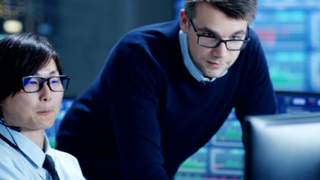 In-the-Network-Operations-Center-Senior-Trader-Teaches-Young-Associate-to-Makes-Personal-Client-Call-with-a-Headset.-They're-Surrounded-by-Monitors-Showing-Data-and-Graphs.