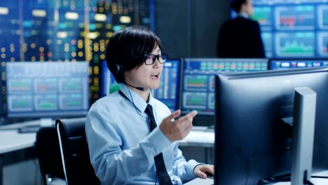 In-the-Network-Operations-Center-Trader-Makes-Personal-Client-Call-with-a-Headset.-He's-Surrounded-by-Monitors-Showing-Stocks-Data-and-Graphs.