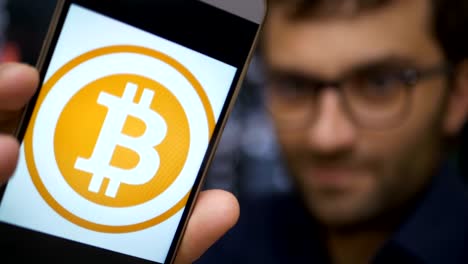 Man-earns-bitcoins-on-his-bitcoin-farm.-Male-making-a-payment-with-bitcoins-cryptocurrency-using-his-smartphone.-Custom-application-interface-design.-Guy-smiles,-earns-on-the-financial-market