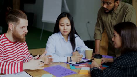 Young-hipster-man-pointing-at-paper-with-plan-of-blockchain-system-implementation-sharing-with-colleagues-sitting-at-the-table-and-discussing-start-up-project