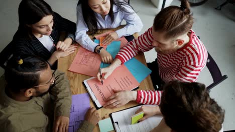 Draufsicht-der-junge-Hipster-männlichen-Anführer-Blockchain-Gründungsvorhaben-mit-Multi-ethnischen-Team-Papierplans-zeigen,-während-sie-am-Tisch-im-gemütlichen-Büro-sitzen-zu-diskutieren