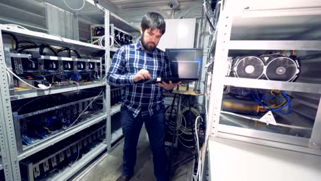 Male-programmer-is-standing-in-the-middle-of-a-mining-farm-unit-with-a-keyboard-and-working