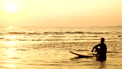 4K.-silhouette-of-surfer-man-relax-by-sitting-on-surfboard-over-the-sea-at-sunset-on-tropical-beach.-sport-and-recreation-for-summer-season-concept