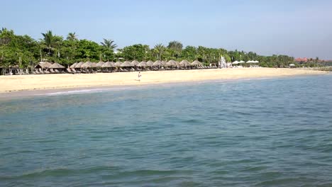 Wellen-des-warmen-Meeres-laufen-am-Sandstrand-des-tropischen-Resorts-mit-Sonnenschirmen-und-Liegestühlen