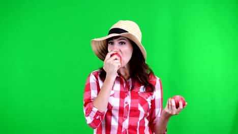 funny,-smiling-female-farmer-in-checkered-shirt-and-hat,-holding-in-hands-two-large-sour-apples,-eats,-bites-them,-wrinkles-her-face,-on-green-background-in-studio.-Healthy-food-to-your-table