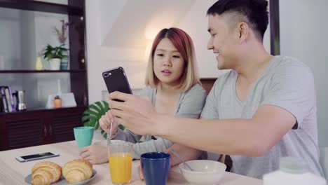 Attractive-young-Asian-couple-distracted-at-table-with-newspaper-and-cell-phone-while-eating-breakfast.-Excited-young-Asian-couple-surprised-by-unbelievably-good-news,-happy-family-amazed-by-internet.