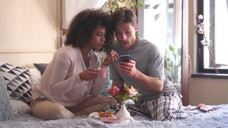 Pareja-desayunando-mientras-usa-el-teléfono-móvil-en-la-cama