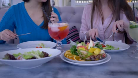 start-diet,-girls-during-a-dinner-communicate-sitting-at-table-with-fork-and-a-knife-eating-Greek-salad-in-cafe