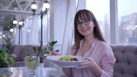 Ernährung,-fröhliches-junges-Mädchen-in-Gläsern-sitzen-im-Café-mit-einer-Gabel-in-der-hand-griechischen-Salat-essen-und-schaut-in-die-Kamera-zu-starten