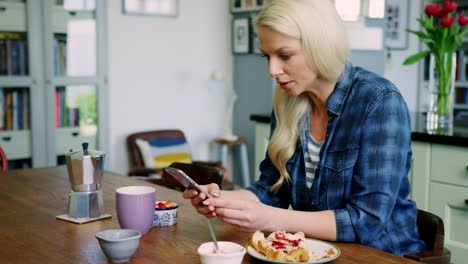 Schöne-blonde-Frau-Überprüfung-Smartphone-am-Frühstückstisch