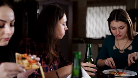 The-company-of-cheerful-girls-in-the-pizzeria.-Girlfriend-in-a-pizzeria-drinking-beer-and-eating-pizza,-chatting-and-having-fun.